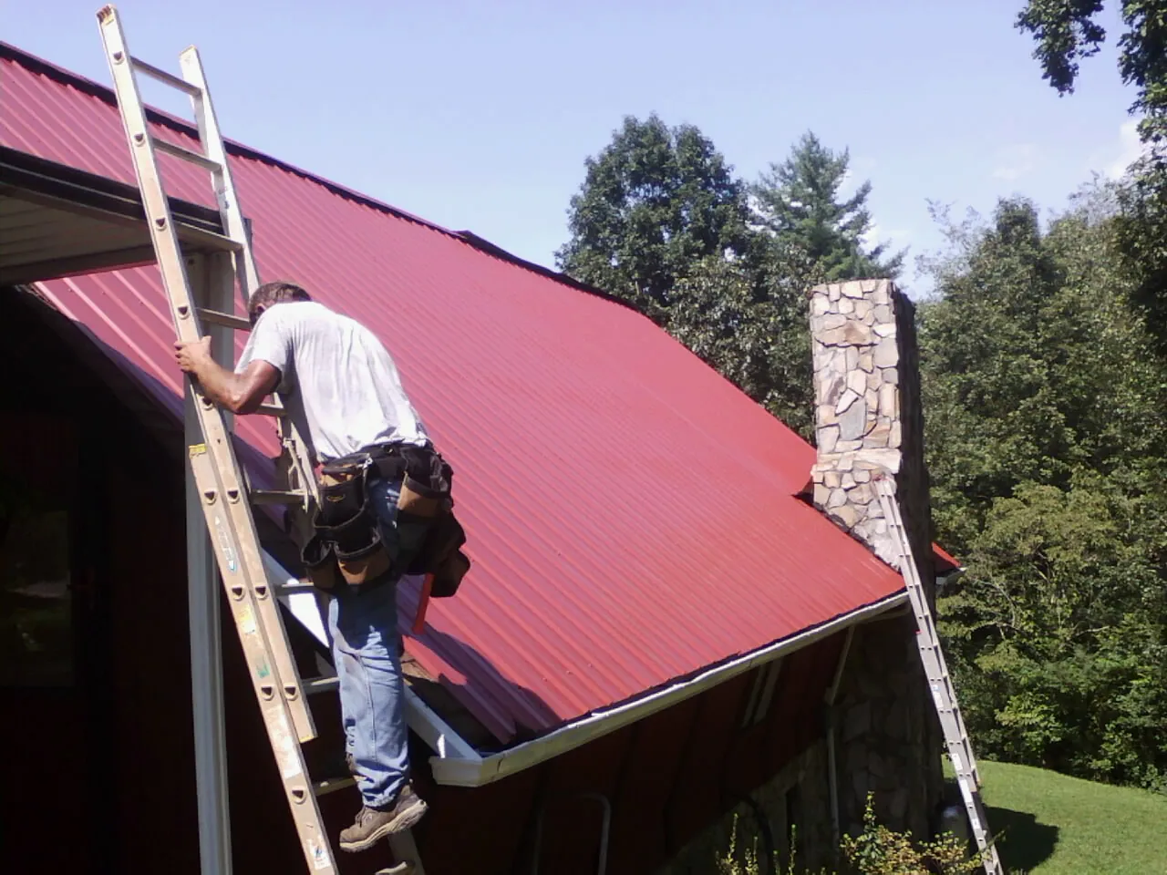 Roofer on a ladder beside a red metal roof and stone chimney — Soffit & Fascia Repair by Roofing Brothers in Glenshaw